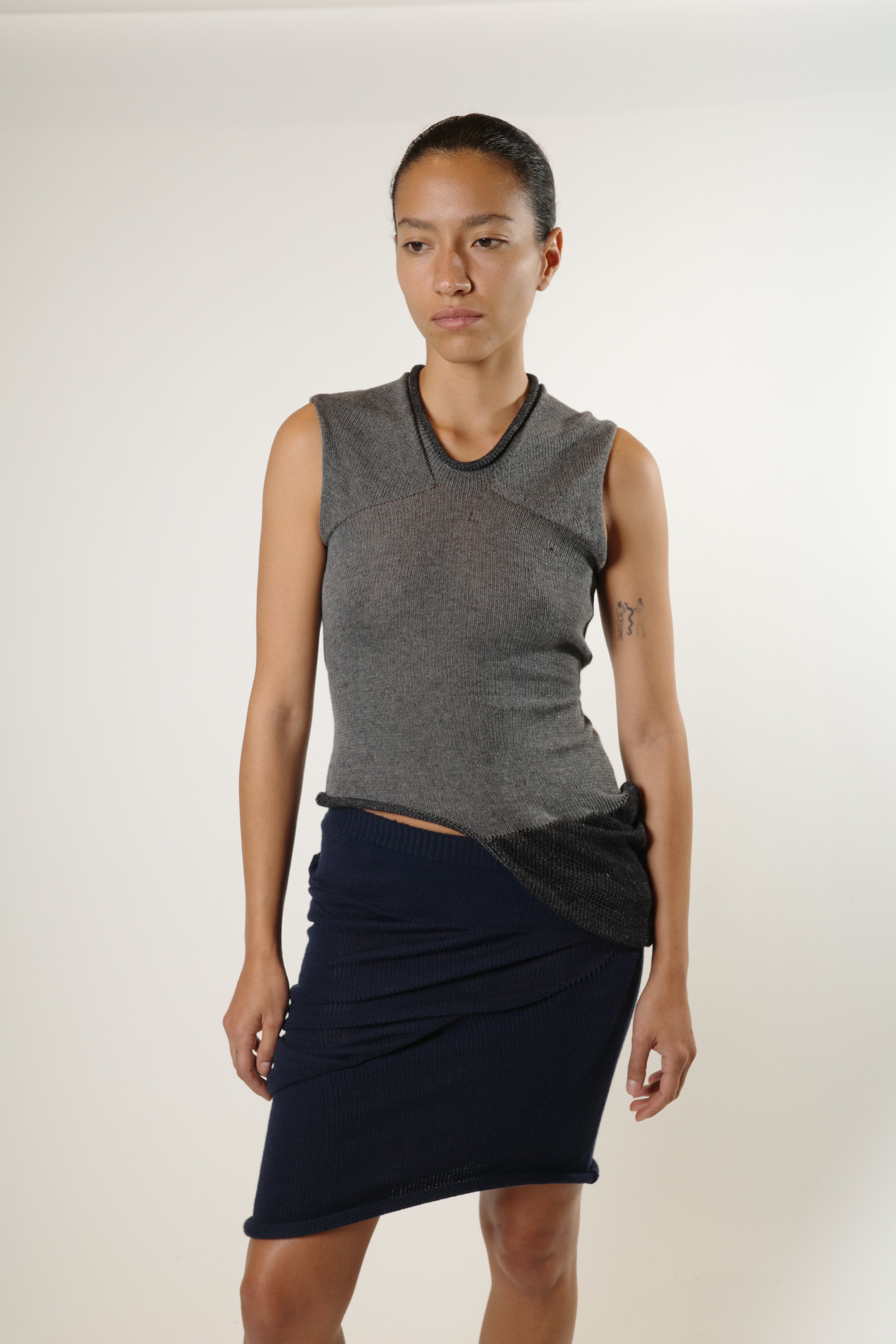 Full body view of grey reversible knit top with draped side detail, styled with navy knit skirt.