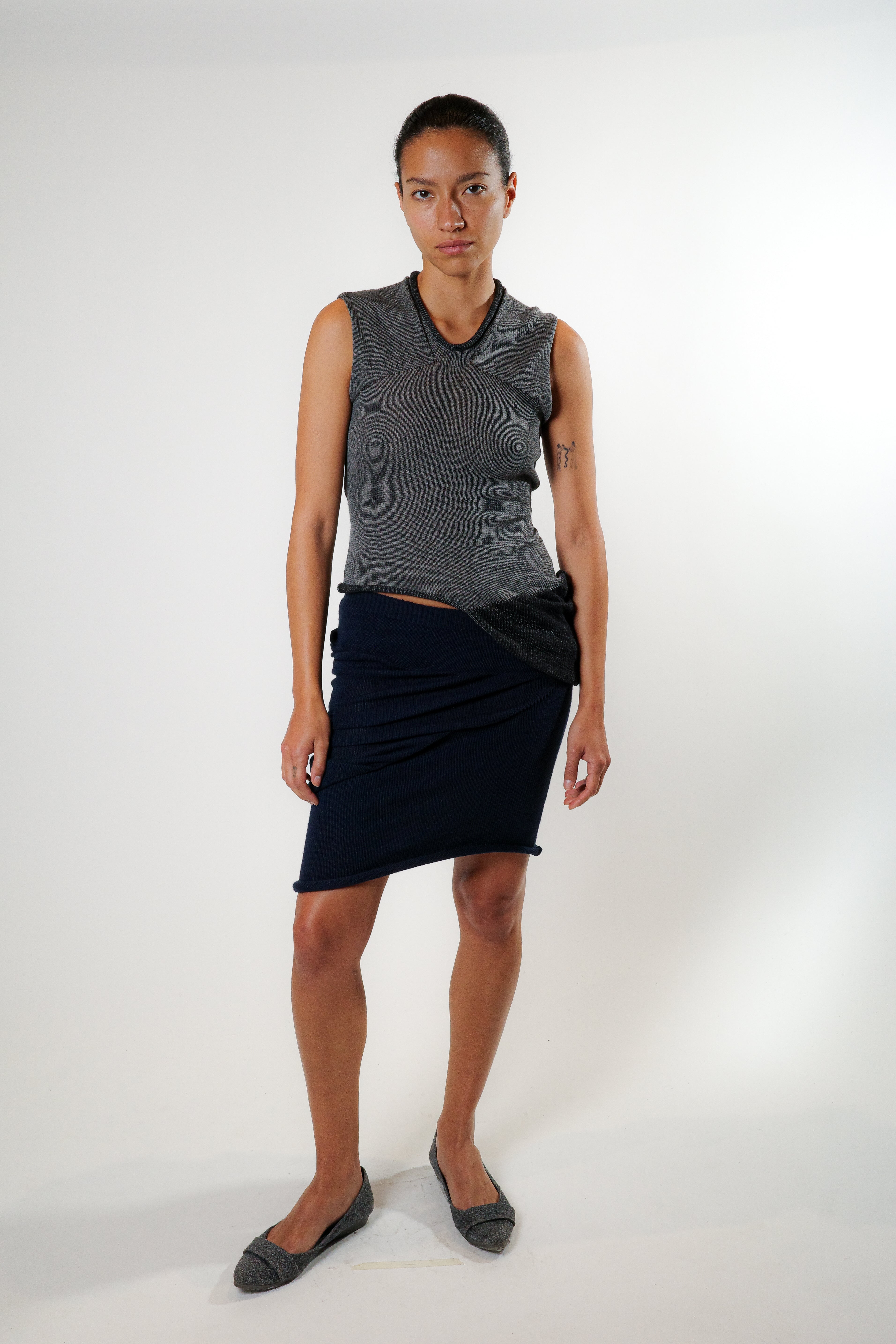Full body view of grey reversible knit top with draped side detail, styled with navy knit skirt.