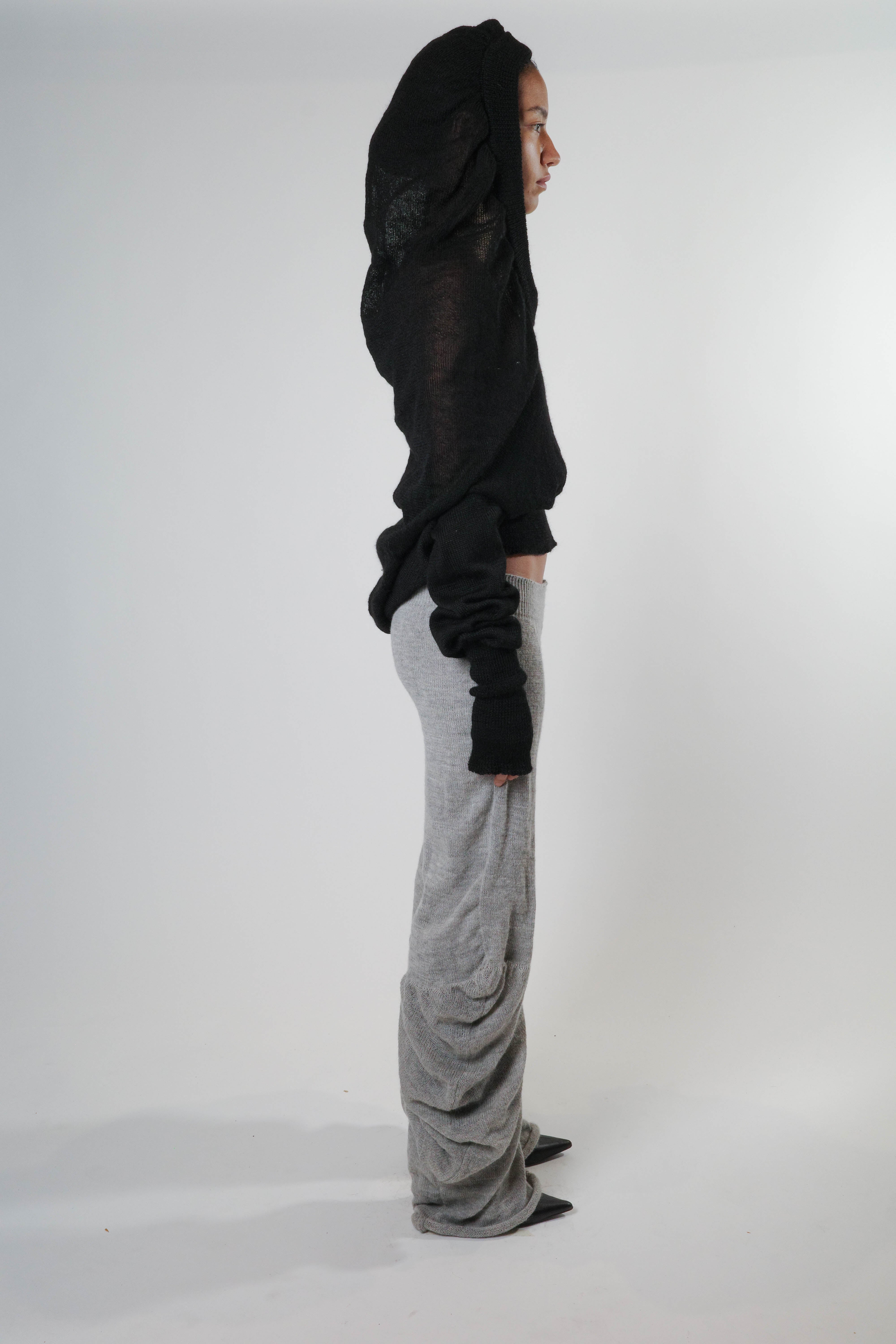Side view of black knit triangle hoodie with ribbed waist and sleeve hems, styled with grey knit pants