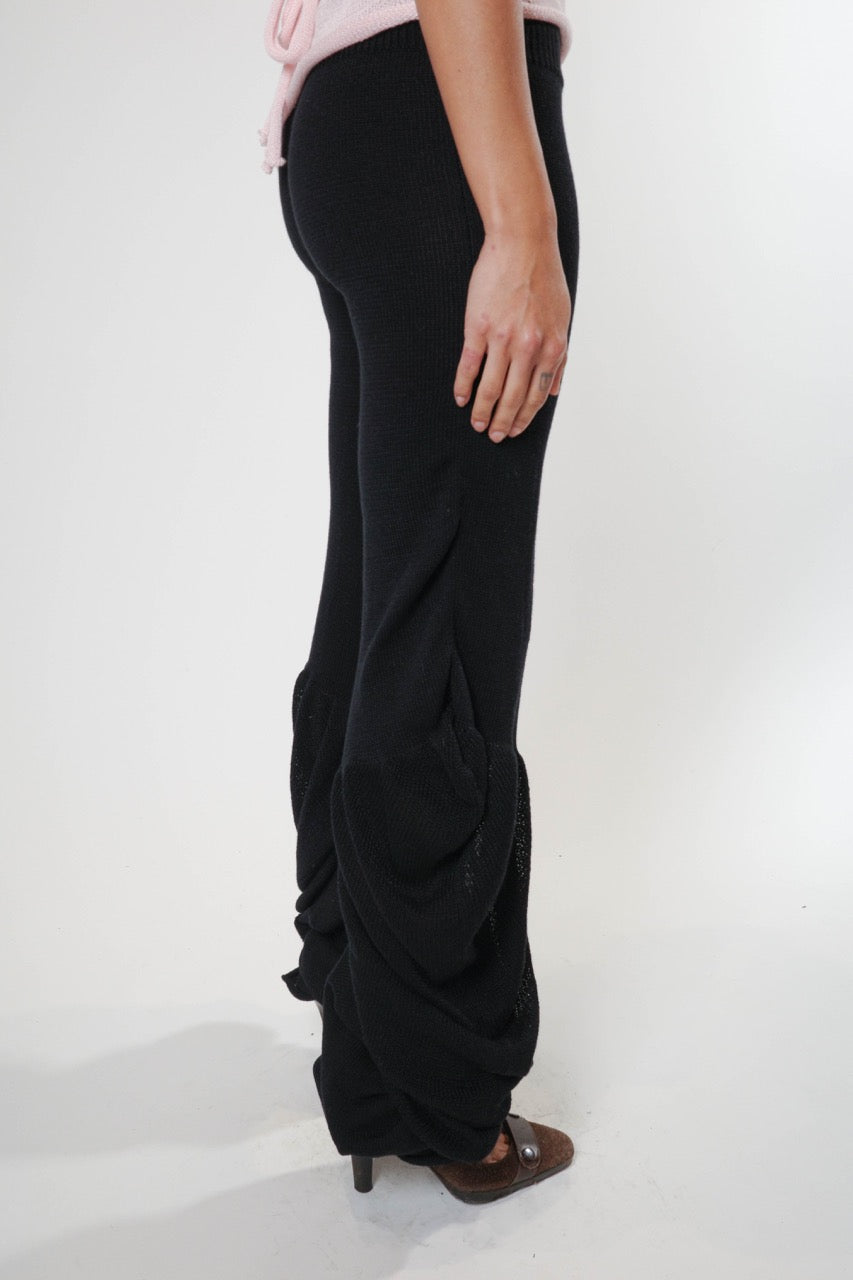 Black knit pants with soft draped detailing along the legs and a fluid, elongated silhouette