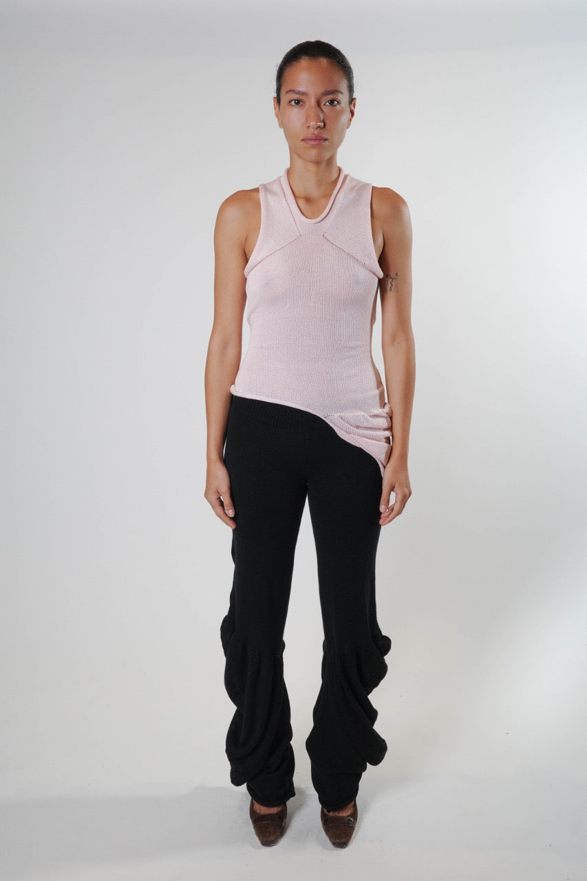 Full body view of black draped knit pants with a fluid, elongated silhouette, styled with a pink knit tank top featuring panel and asymmetric hem detailing.