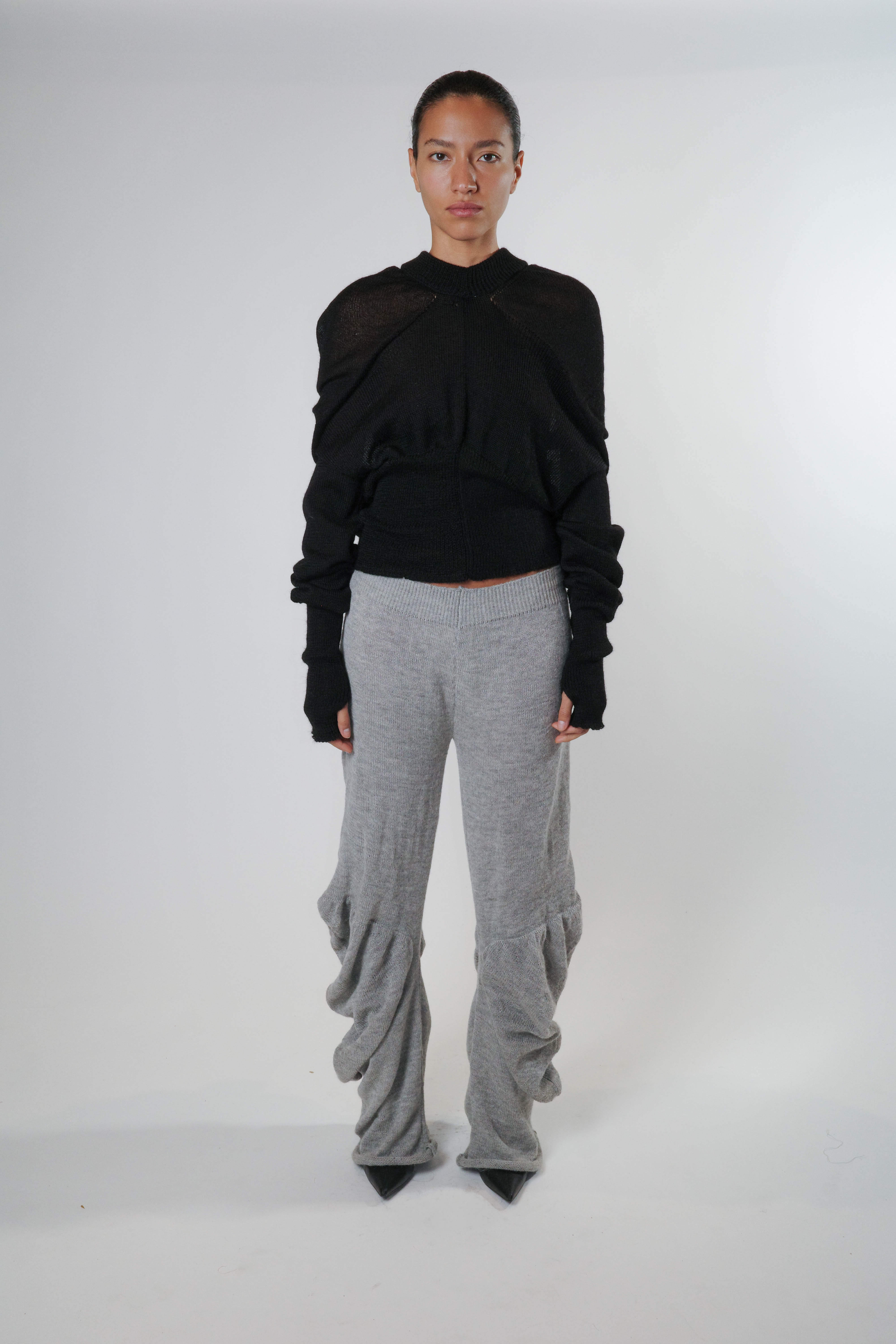 Full body view of black knit triangle hoodie with ribbed waist and cuffs, paired with grey knit pants